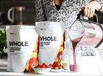 Vivo Life Whole Plant-Based Nutritional Shake supplement in a glass with leafy greens and scoops of powder on a wooden table.