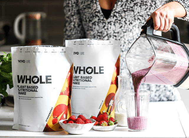 Vivo Life Whole Plant-Based Nutritional Shake supplement in a glass with leafy greens and scoops of powder on a wooden table.